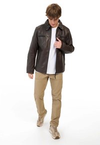 Donkerbruin leren jack met een kraagontwerp, voorzakken met rits en een gladde textuur. Gedragen over een wit shirt en gecombineerd met beige broek.