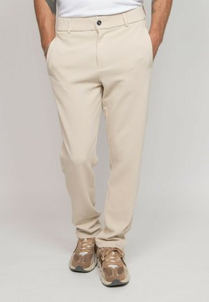 Homme portant un pantalon beige sur mesure et des baskets marron, les mains dans les poches, debout devant un fond clair uni.