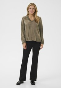Olivenfarvet bluse med lange ærmer, tekstureret ruffled V-hals og elastiske manchetter, parret med sorte flared bukser og sorte sko.