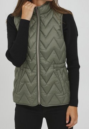 Kvinde iført olivengrøn quiltet ærmeløs vest med lynlås over sort langærmet top, hånd rører ved kraven, stående mod en ensfarvet baggrund.