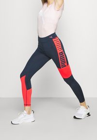 Leggings blu navy con pannelli rossi a contrasto, caratterizzati da un audace design del logo laterale. Tessuto liscio e elastico, ideale per l'abbigliamento sportivo.