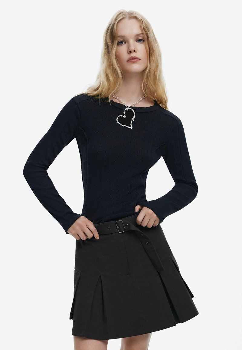 Tengeri kék, hosszú ujjú, bordázott pulóver, fekete, pliszírozott szoknyával, övvel, amely textúrázott felületű, valamint ezüst szív alakú nyaklánccal kombinálva.