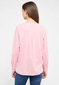 Frau mit langen braunen Haaren, die eine rosa Bluse mit langen Ärmeln und kleinen weißen Punkten trägt, kombiniert mit blauen Jeans, mit dem Rücken zur Kamera vor einem weißen Hintergrund.