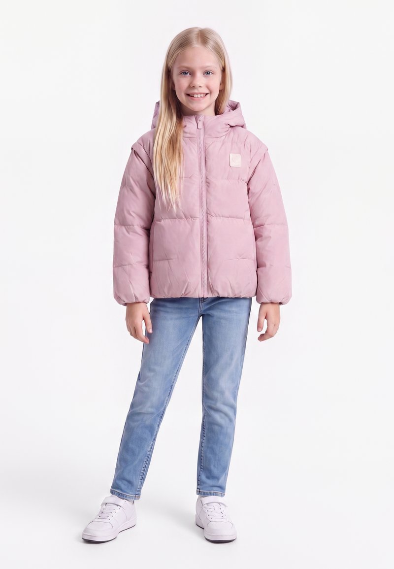 Jeune fille aux longs cheveux blonds portant une doudoune rose clair, un jean bleu clair et des baskets blanches, debout et souriante.