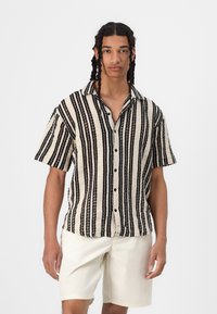 Camisa de manga corta en rayas crema y negra, con botón frontal y tejido texturizado, combinada con pantalones cortos de color claro.