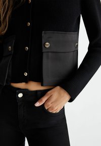 Kvinde iført sort ribstrikket cardigan med store klaplommer og sorte bukser, hånden hvilende i forlommen mod en lys baggrund.