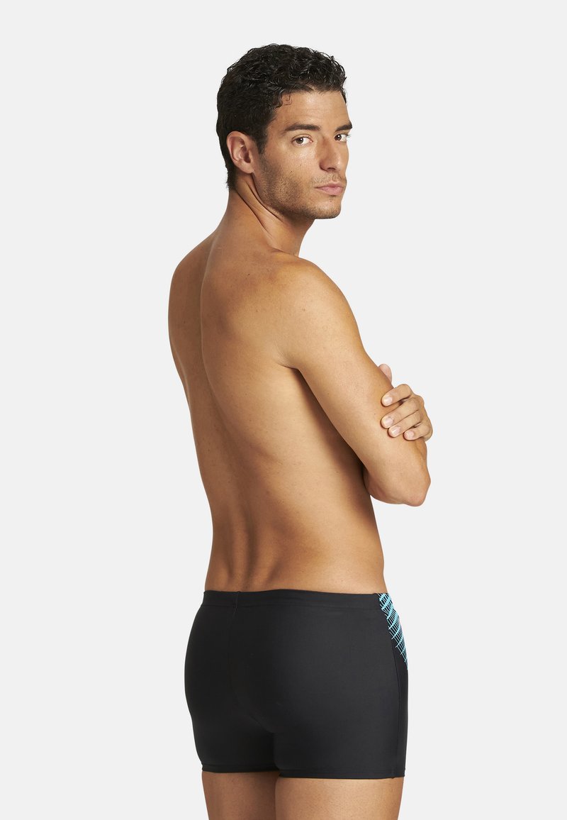 Mężczyzna bez koszulki w czarnych kąpielówkach z niebieskim wzorem, stojący odwrócony plecami z założonymi rękami, spoglądający przez ramię na białym tle.