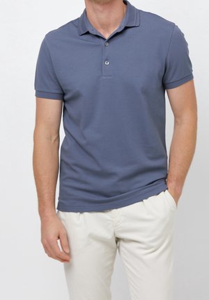 Homme portant un polo bleu à manches courtes et un pantalon blanc, debout avec une main dans la poche devant un fond uni.