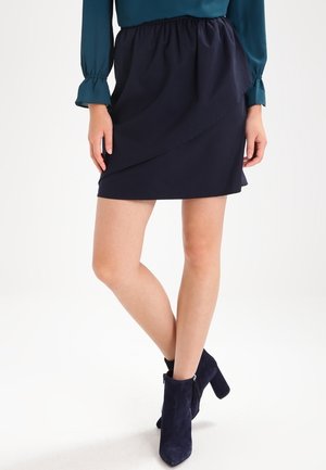Femme portant une jupe bleu foncé associée à des bottines assorties et un chemisier manches longues de couleur sarcelle, debout, jambes croisées.