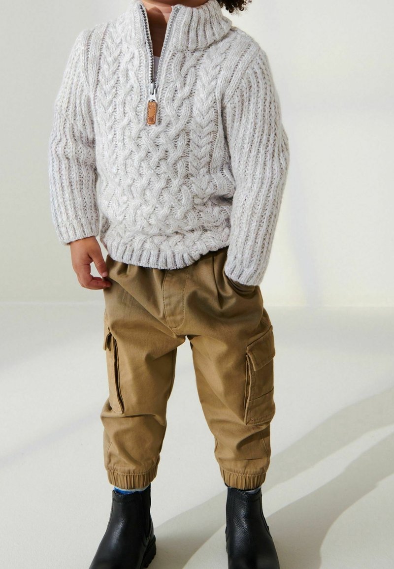 Enfant portant un pull gris clair à torsades avec une demi-fermeture éclair et un pantalon cargo marron avec des poignets élastiques, debout les mains dans les poches.