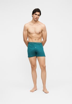 Hemdloser Mann steht barfuß und trägt türkisfarbene Calvin Klein Boxershorts mit Knopfleiste vor einem einfarbigen weißen Hintergrund.