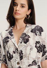 Kobieta z długimi ciemnymi włosami, nosząca złote kolczyki w kształcie kół oraz beżową koszulę w kwiatowy wzór, na której znajdują się czarno-białe motywy kwiatowe.