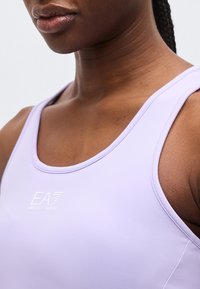 Nærbilde av en person som har på seg en lys lilla sportstopp med EA7 Emporio Armani-logoen sentrert på brystet.
