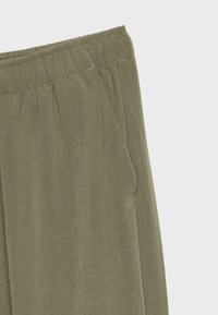 Gros plan de la taille d'un pantalon vert olive avec bande élastique et détail visible d'une poche latérale sur fond clair.