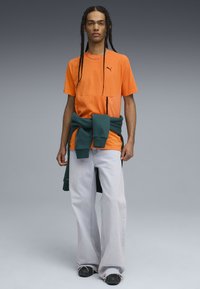 Ung mand med lange fletninger har en orange Puma T-shirt, lyse brede bukser, sorte sneakers og en mørkegrøn sweatshirt bundet om livet.