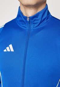 Kék sportdzseki magas gallérral, cipzáras záródással, fehér részletekkel, és jól látható márkajellel a bal mellkas tájékán. Puha, texturált anyag.