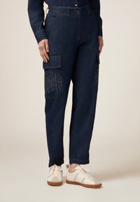Pantaloni cargo in denim blu scuro con tasche laterali, caratterizzati da cuciture verticali decorative. Abbinati a sneakers beige.