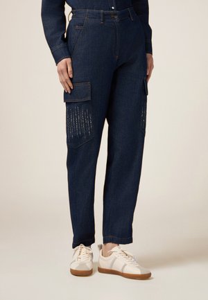 Pantalones cargo de mezclilla azul oscuro con bolsillos laterales, que presentan costuras decorativas verticales. Combinados con zapatillas beige.