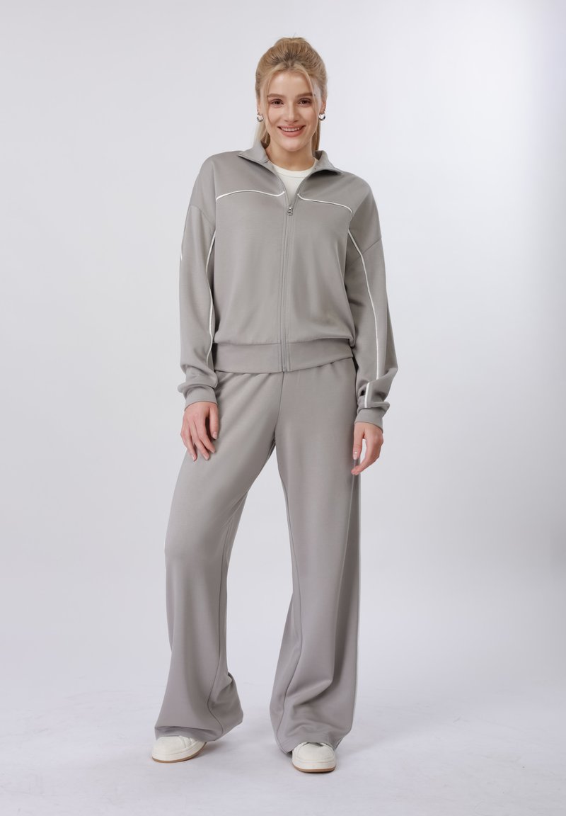 Jeune femme souriante, portant un survêtement gris clair zippé avec des passepoils blancs et un pantalon à jambes larges, debout devant un fond clair uni.