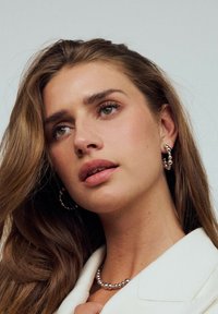 Silverpärlade örhängen och ett matchande halsband, som bärs av en kvinna med långt hår, mot en ljus enfärgad bakgrund.