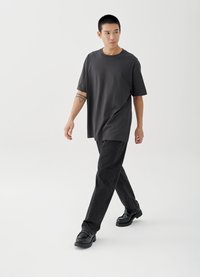 Pilka didelio dydžio marškinėliai derinami su juodais džinsais ir juodais batais. Audinys atrodo minkštas, lygaus paviršiaus ir laisvo kirpimo.