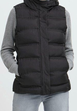 Personne portant un gilet matelassé noir sur un pull en maille gris, les mains dans les poches du gilet et un jean bleu clair.