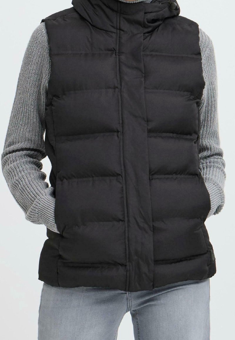 Personne portant un gilet matelassé noir sur un pull en maille gris, les mains dans les poches du gilet et un jean bleu clair.