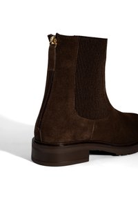 Bottine en suède marron avec des panneaux latéraux en élastique ribbé, une fermeture éclair avec des garnitures dorées et une semelle en caoutchouc texturée.