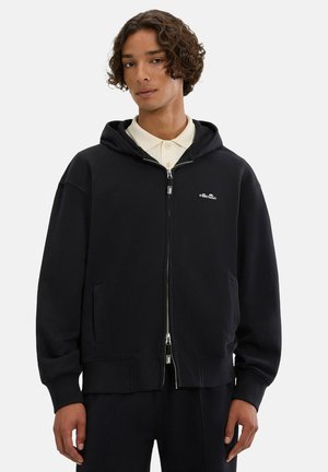 Junge Person mit lockigem Haar, trägt einen schwarzen Reißverschluss-Hoodie über einem beigen Hemd mit Kragen und steht vor einem einfarbigen Hintergrund.