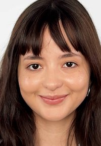 Portrait en gros plan avec des cheveux bruns, lisses et une frange droite. Le visage a un teint lisse, portant de petites boucles d'oreilles en argent.