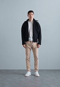 Veste zippée noire sur un t-shirt blanc, pantalon beige et sneakers blanches ; la veste possède un col haut et des poignets côtelés.
