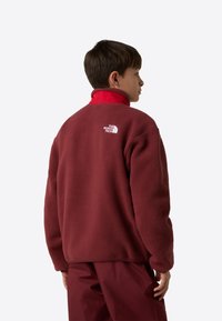 Μπουφάν από φλις μπορντό με ψηλό κόκκινο γιακά, με λευκό λογότυπο στο πίσω μέρος. Μαλακή υφή και άνετη εφαρμογή, μακριά μανίκια.