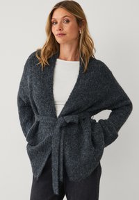 Frau trägt eine dunkelgraue strukturierte Strickjacke mit Gürtel über einem weißen Hemd und schwarzen Hosen, Hände in den Taschen, steht vor einem schlichten Hintergrund.