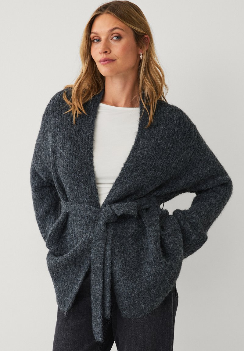 Frau trägt eine dunkelgraue strukturierte Strickjacke mit Gürtel über einem weißen Hemd und schwarzen Hosen, Hände in den Taschen, steht vor einem schlichten Hintergrund.