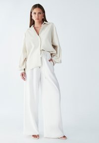 Camicia oversized beige chiaro con bottoni e maniche lunghe, abbinata a pantaloni bianchi a gamba larga con vita regolabile e un sottile accento dorato.