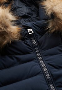 Navy blue puffer jacket with a detachable beige fur hood, featuring a prominent black zipper and a silver logo tag near the collar.