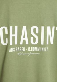 Žalias medvilninis marškinėliai su ryškiu baltu tekstu: "CHASIN' AMS BASED - C.COMMUNITY" su papildomu mažesniu tekstu žemiau. Lygus paviršius.