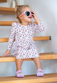 Blumenmusterkleid in hellrosa mit langen Ärmeln, kombiniert mit pinken herzförmigen Sonnenbrillen und lila Schuhen, sitzt auf Holzstufen.