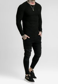 Schwarzes, geripptes Langarmoberteil kombiniert mit kaputten schwarzen Jeans und schwarzen Schuhen mit goldenen Akzenten. Texturierte Materialien, figurbetontes Design.