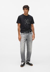 Mladý muž stojaci v čiernom grafickom tričku, vyblednutých šedých džínsach, čiernom opasku a čiernych teniskách s bielymi šnúrkami na bielom pozadí.