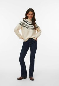 Cremfarbener Strickpullover mit schwarzem geometrischem Muster an der Schulterpartie, kombiniert mit dunkelblauen, ausgestellten Jeans und braunen Stiefeletten.