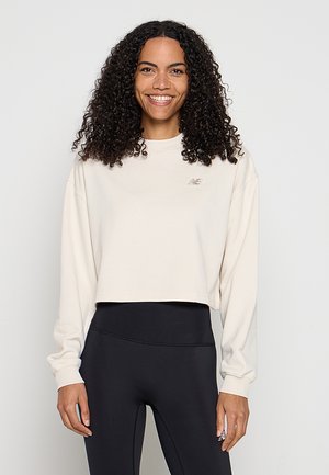 Lächelnde Frau mit lockigem Haar, die ein cremefarbenes, bauchfreies Sweatshirt und schwarze Leggings mit hoher Taille trägt, vor einem schlichten Hintergrund stehend.