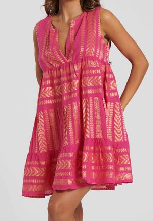 Robe rose sans manches avec des motifs géométriques dorés, coupe ample, col en V et jupe à volants, portée par une personne avec une main sur la hanche.