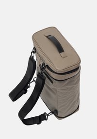 Beige laukku, jossa on suorakaiteen muotoinen muoto, yläkahva, säädettävät mustat hihnat ja vetoketju sulkemista varten. Valmistettu teksturoidusta materiaalista.