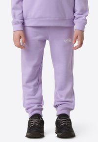 Lila Jogginghose aus weichem Material mit elastischen Bündchen und einem kleinen weißen Logo. Kombiniert mit schwarzen Sportschuhen mit strukturierter Sohle.