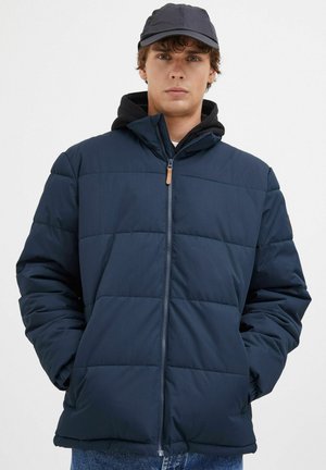 Marineblå pufferjakke med quiltet design, lynlåslukning og hætte. Bæres over en sort hoodie, med diskret læderdetalje på lynlåsen.