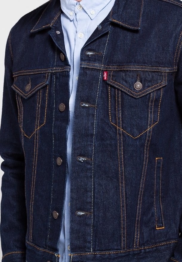 Veste en denim bleu foncé avec des surpiqûres orange contrastantes, portée sur une chemise boutonnée bleu clair, avec des poches à rabat sur la poitrine et des boutons en métal.