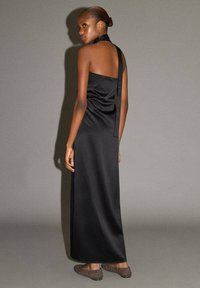 Robe noire en satin avec un col licou et un dos nu, silhouette longue et fluide. Le mannequin porte des chaussures marron à enfiler avec des accents cloutés.