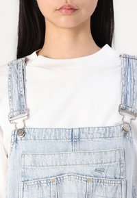 Salopette en denim bleu clair avec des clips en métal ajustables, des poches avant et un t-shirt blanc à col rond en dessous. Design simple et décontracté.