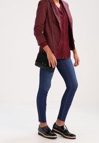 Donna in giacca bordeaux e blusa, jeans skinny blu, scarpe in pelle verniciata nera, che tiene una piccola borsa nera con tracolla a catena.